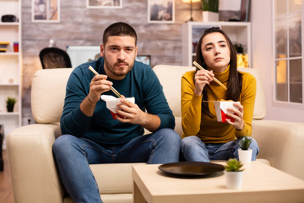 Na moderna e acolhedora sala de estar casal está desfrutando de macarrão takeaway enquanto assiste TV
 - Foto, Imagem