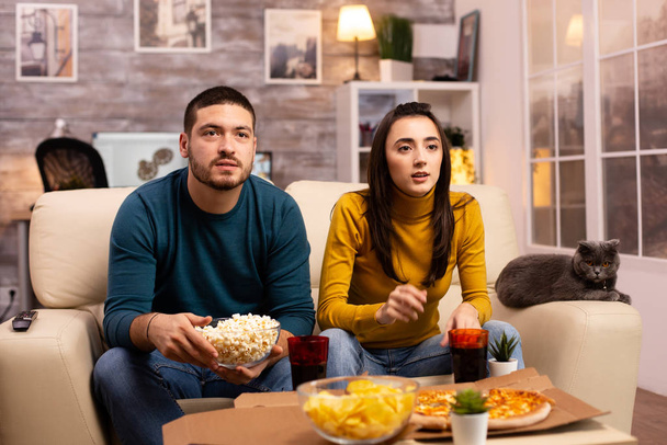Beautiful young couple watching TV and eating fast food takeaway - Photo, Image