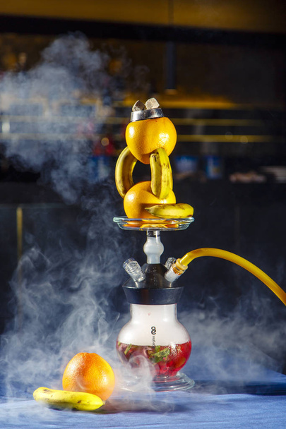Hookah on oranges and bananas. Citrus hookah. Hookah with round clear bulb with a red liquor. A bowl of hookah made of oranges, in which bananas are inserted.  - Фото, зображення