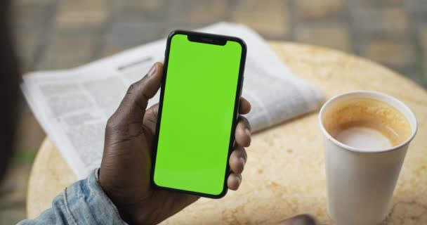 Lviv, Ukraine - August 09, 2019: Hands of Afro American Man Sitting at Table with News Paper and Coffee Cup on it, Holding Smartphone with Green Screen and Drinking Coffee - Filmmaterial, Video