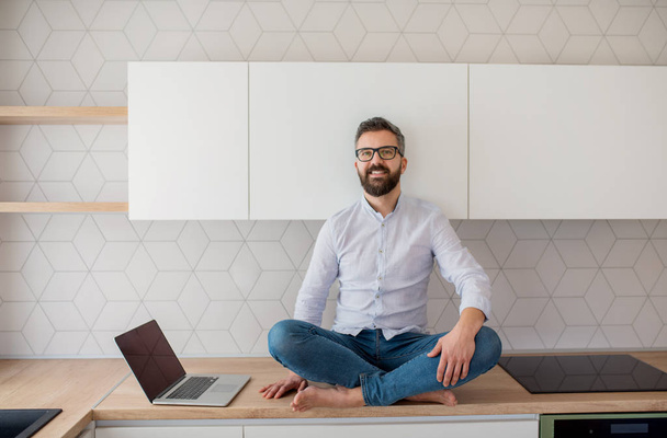 A mature man with laptop sitting in kitchen in unfurnished new house. - Fotoğraf, Görsel