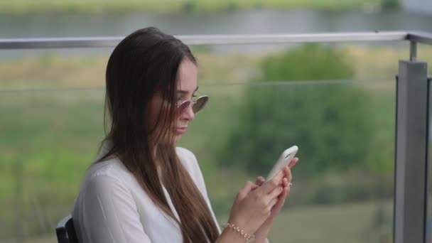 νεαρή γυναίκα δακτυλογραφεί sms στο smartphone κάθεται στη βεράντα το καλοκαίρι - Πλάνα, βίντεο