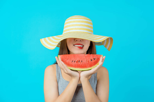 Азиатская молодая женщина в летней шляпе и с горкой арбуза на изолированном голубом цветовом фоне, Сексуальный укус рта, пространство для копирования и студия, модные путешествия и туристическая концепция - Фото, изображение