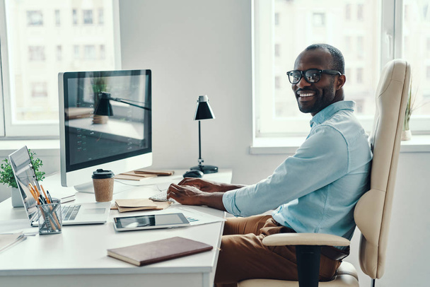 Happy young African man in shirt using computer and smiling while working in the office    - Fotó, kép