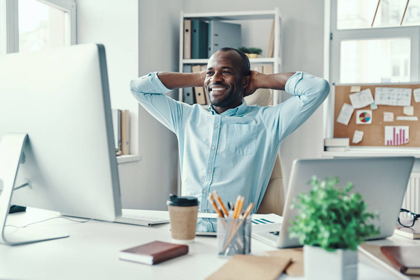 Handsome young African man in shirt keeping hands behind head and smiling while working in the office     - Фото, изображение