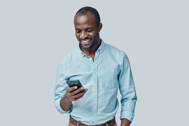 Handsome young African man using smartphone and smiling while standing against grey background      - Foto, immagini
