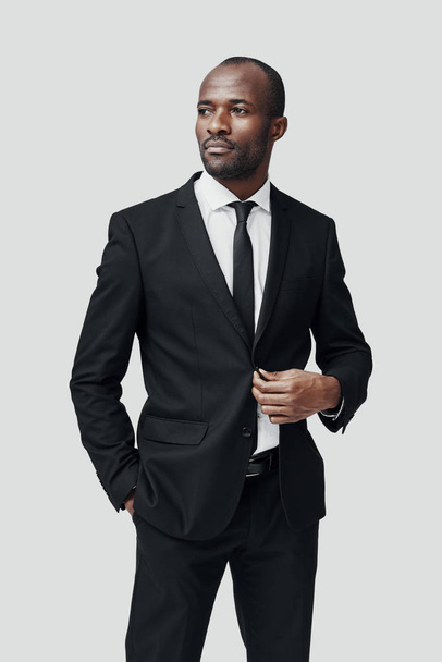 Stylish young African man in formal wear looking away and adjusting jacket while standing against grey background               - Foto, imagen