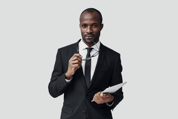 Charmanter junger afrikanischer Mann in formeller Kleidung, der mit einem digitalen Tablet arbeitet, während er vor grauem Hintergrund steht - Foto, Bild