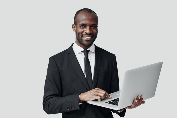 Confident young African man in formal wear working using computer while standing against grey background  - Photo, image