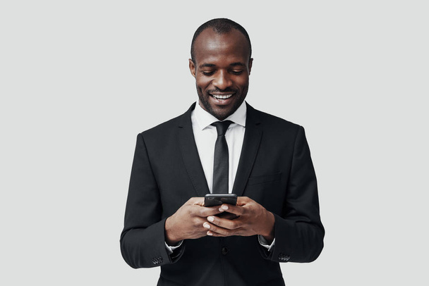 Modern young African man in formal wear using smartphone and smiling while standing against grey background                  - Fotó, kép