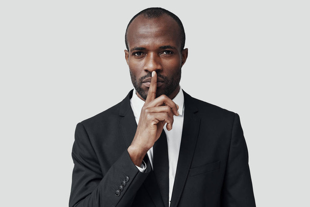 Hombre africano joven misterioso en desgaste formal manteniendo el dedo en los labios mientras está de pie sobre fondo gris
 - Foto, imagen