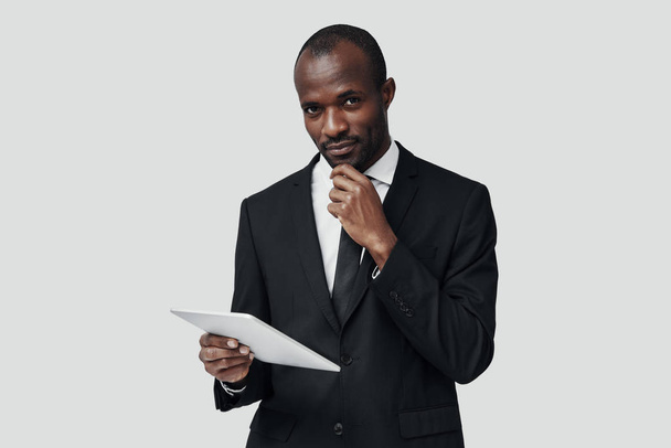 Successful young African man in formal wear working using digital tablet while standing against grey background     - Фото, изображение