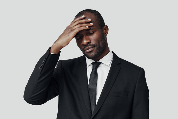 Tired young African man in formal wear suffering from headache while standing against grey background  - Fotoğraf, Görsel
