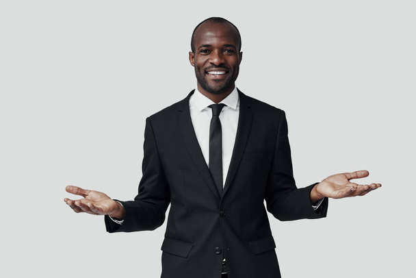 Elegante jonge Afrikaanse man in formele kleding die naar de camera kijkt en glimlacht terwijl hij tegen een grijze achtergrond staat - Foto, afbeelding