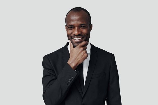 Stylish young African man in formal wear looking at camera and smiling while standing against grey background - Foto, Imagem