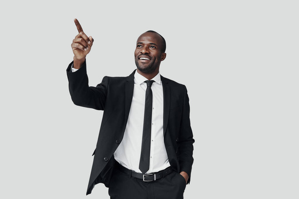 Handsome young African man in formal wear pointing copy space and smiling while standing against grey background - Photo, image