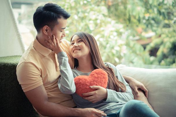 Kanepede oturan Asyalı çiftler kırmızı bir kalbi tutan ve mutlu bir şekilde gülümseyen kadınlar. Aşıklar için sevgi ve samimiyet kavramı, Sevgililer Günü. - Fotoğraf, Görsel