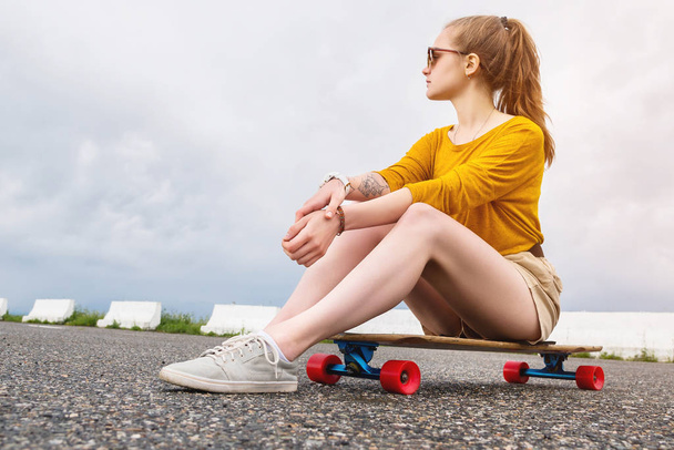 Attractive young girl with a tattoo on her arm in sunglasses and shorts sits on her longboard in a suburban parking on a background of gray sky. Millennials generation and leisure concept - Photo, image