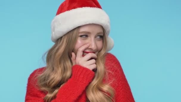 Close up view of Playful shy pretty woman in christmas hat embarrassing and covering her mouth over turquoise background - Footage, Video