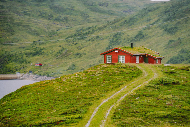 Norweski drewniany domek letni z widokiem na malownicze jezioro, Norwegia, Skandynawia. Domek nad jeziorem na wsi. Chata pokryta torfem nad jeziorem. Typowa chata pokryta trawą w Norwegii. Typowa czerwona chatka rybacka rorbu - Zdjęcie, obraz