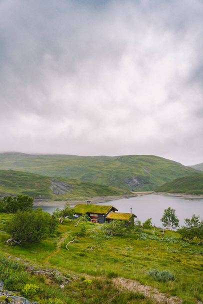 Norweski drewniany domek letni z widokiem na malownicze jezioro, Norwegia, Skandynawia. Domek nad jeziorem na wsi. Chata pokryta torfem nad jeziorem. Typowa chata pokryta trawą w Norwegii. Typowa czerwona chatka rybacka rorbu - Zdjęcie, obraz