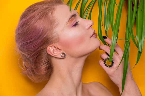 Shiny golden make-up on a beautiful young European girl model and leaves of a green plant isolated on an orange background in a photo studio - Фото, изображение