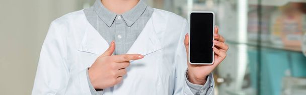 Vista ritagliata del farmacista che punta con il dito lo smartphone con schermo bianco, colpo panoramico
 - Foto, immagini