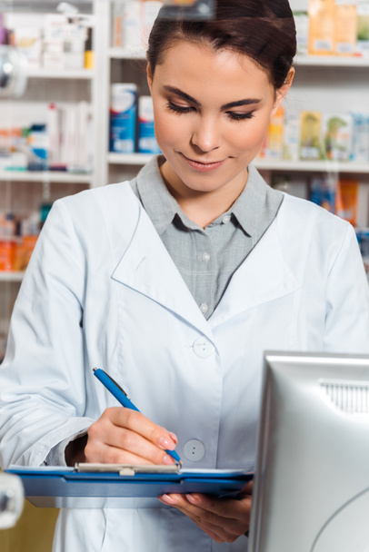 Attractive pharmacist writing on clipboard at pharmacy counter - Φωτογραφία, εικόνα