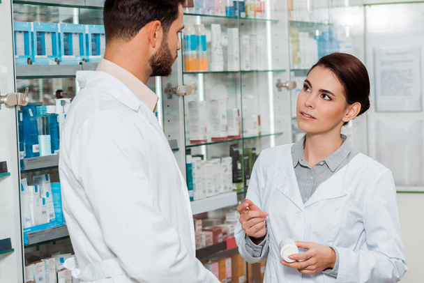 Pharmacist with jar of pills looking at colleague in pharmacy - 写真・画像