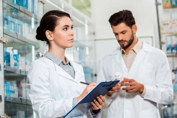 Pensive pharmacist writing on clipboard and colleague with pills on background - Photo, Image