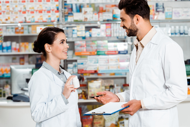 Vista lateral de farmacêuticos com pílulas e prancheta sorrindo uns para os outros na farmácia
 - Foto, Imagem