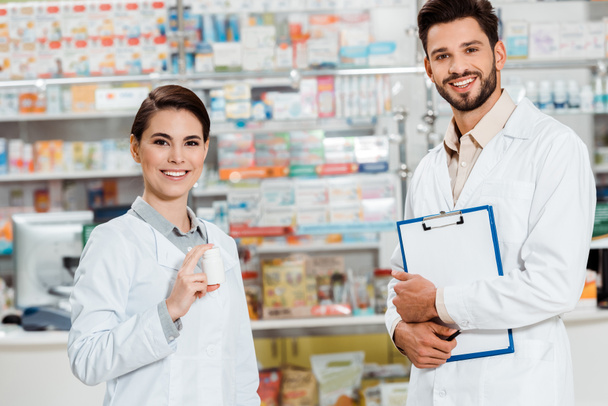 Farmacéuticos sonrientes con portapapeles y pastillas mirando la cámara en la farmacia
 - Foto, Imagen