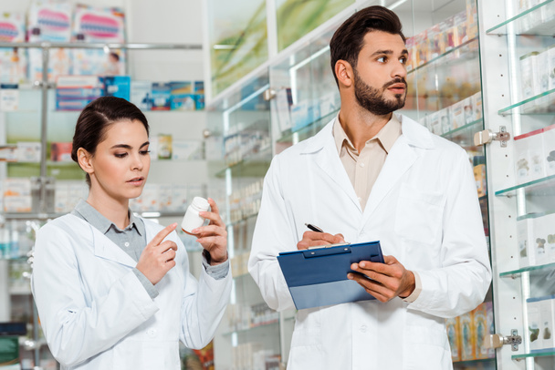 Farmacéutico con portapapeles y colega con pastillas por escaparate de farmacia
 - Foto, Imagen