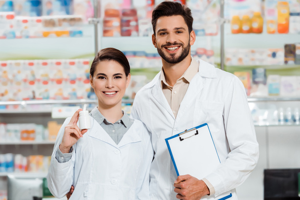 Farmacéutico sonriente con portapapeles y frasco de pastillas mirando a la cámara en la farmacia
 - Foto, Imagen