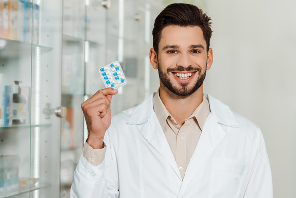 Όμορφος φαρμακοποιός χαμογελά στην κάμερα, ενώ κρατώντας κυψέλη με χάπια  - Φωτογραφία, εικόνα