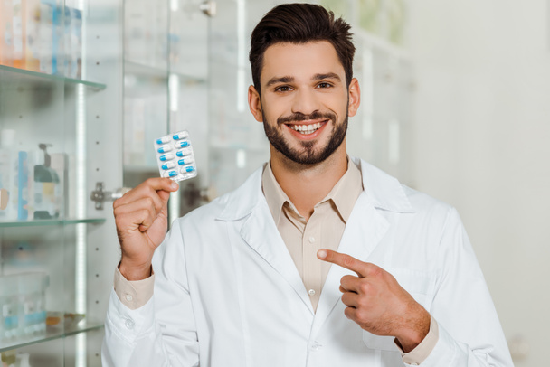 Beau pharmacien souriant à la caméra et pointant vers blister avec des pilules
 - Photo, image