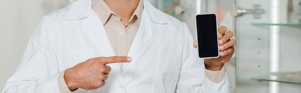 Vista recortada del farmacéutico en bata blanca apuntando al teléfono inteligente con pantalla en blanco, plano panorámico
 - Foto, Imagen