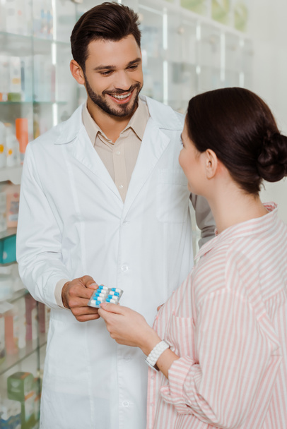 Smiling druggist giving blister with pills to customer by pharmacy showcase - Foto, imagen