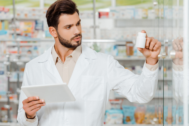 Beau pharmacien tenant tablette numérique et regardant pot de pilules
 - Photo, image