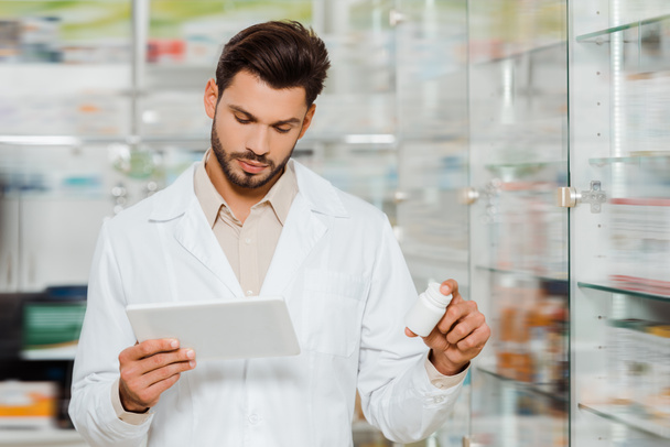 Guapo farmacéutico usando tableta digital mientras sostiene pastillas en la farmacia
 - Foto, imagen