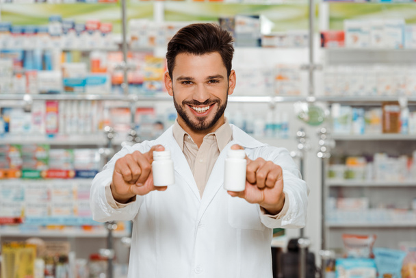 Χαμογελώντας φαρμακοποιός κοιτάζοντας κάμερα και κρατώντας βάζα με χάπια - Φωτογραφία, εικόνα