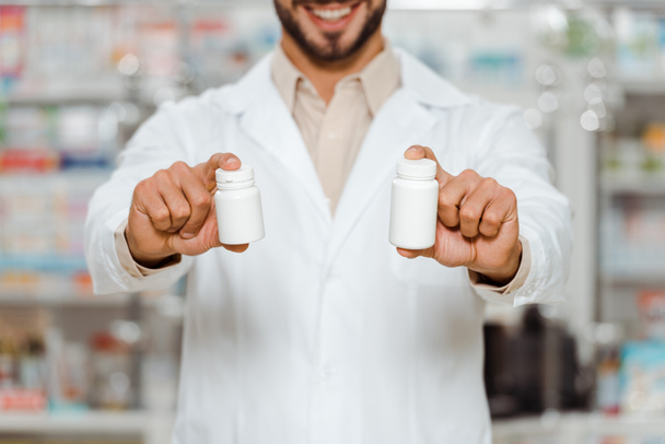 Viljelty näkymä hymyilevä farmaseutti osoittaa purkit pillereitä, valikoiva keskittyä
 - Valokuva, kuva