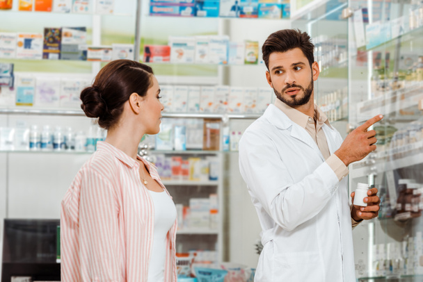 Druggist with pills pointing with finger at shelves with medicaments to customer - Foto, Imagem