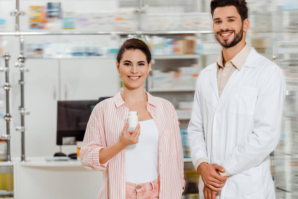Smiling woman with jar of pills beside pharmacist with drugstore showcase at background - Foto, Bild