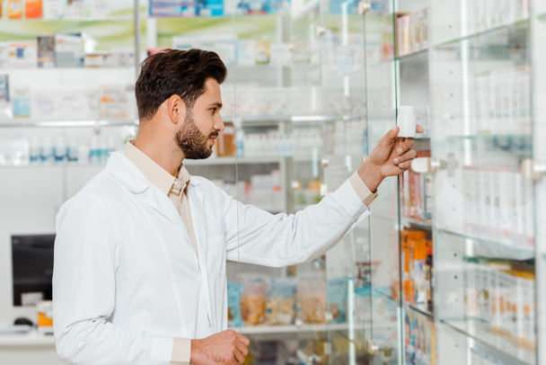 Vista lateral del guapo farmacéutico mirando frasco con pastillas al lado de estantes con medicamentos
 - Foto, Imagen