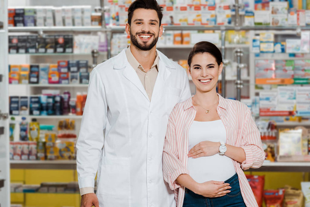 Mosolygós gyógyszerész és terhes nő néz kamera gyógyszertár kirakat a háttérben - Fotó, kép