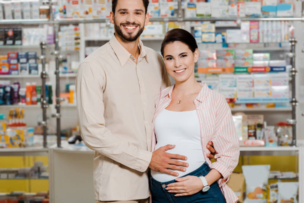 Uomo sorridente che abbraccia moglie incinta e guarda la fotocamera in farmacia
  - Foto, immagini