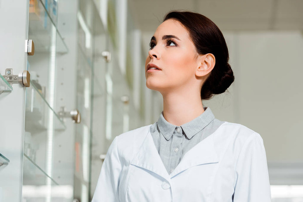 Low angle view of attractive pharmacist looking at medicaments in drugstore showcase - Photo, Image