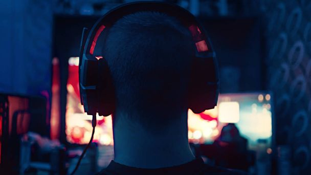 a gamer is sitting in front of a large monitor with headphones,a silhouette in headphones against the background of a monitor with a computer game.photo taken in a dark key in neon light. guy plays computer games at home - Фото, зображення