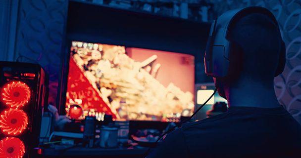 геймер сидит перед большим монитором с наушниками, силуэт в наушниках на фоне монитора с компьютерной игрой. парень играет в компьютерные игры дома
 - Фото, изображение